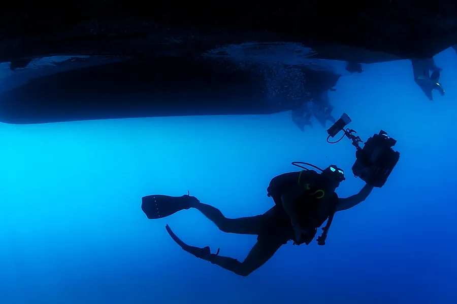 Buzo con equipo de buceo trabajando bajo el casco oscuro de una embarcación, con la luz del agua al fondo.